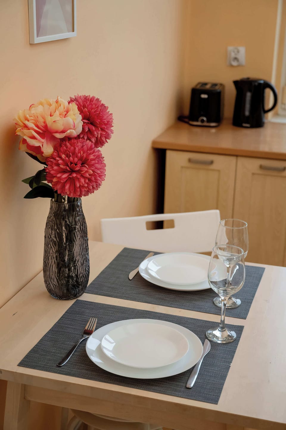A small dining table set with plates and a flower arrangement. The setting feels simple and ready for use. Warm tones add a cozy atmosphere.