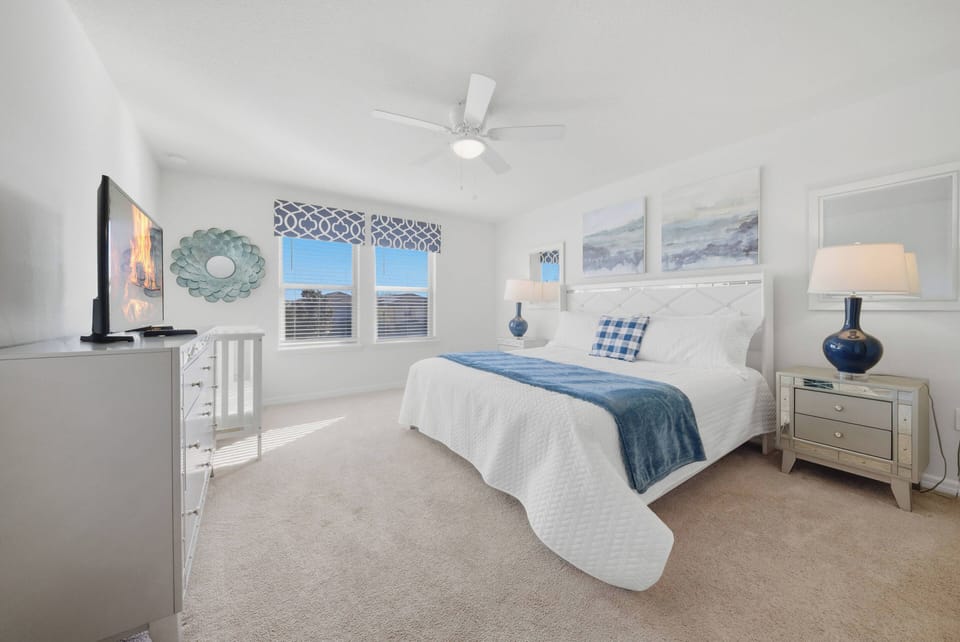 This cozy bedroom features a plush bed with crisp white linens and a soft blue throw for restful nights. Large windows flood the room with natural light, enhanced by a modern dresser and stylish lamps for an inviting atmosphere.