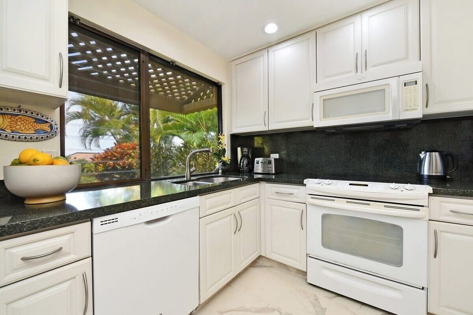 A bright kitchen with white cabinetry, dark granite counters, and garden views through wide windows.