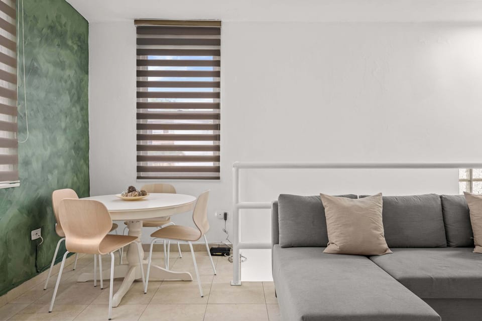 Espacio de concepto abierto. En primer plano, sofá seccional gris junto a una barandilla blanca. Al fondo, mesa de comedor redonda blanca frente a una pared verde. Dos ventanas con persianas y unidad de aire acondicionado instalada en la pared.