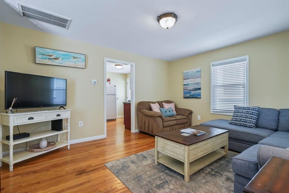 Welcome to this bright and airy living area, combining the warmth of hardwood floors with comfortable, modern seating for a truly inviting space. The stylish gray sectional and unique distressed coffee table are perfect for gathering to watch TV 