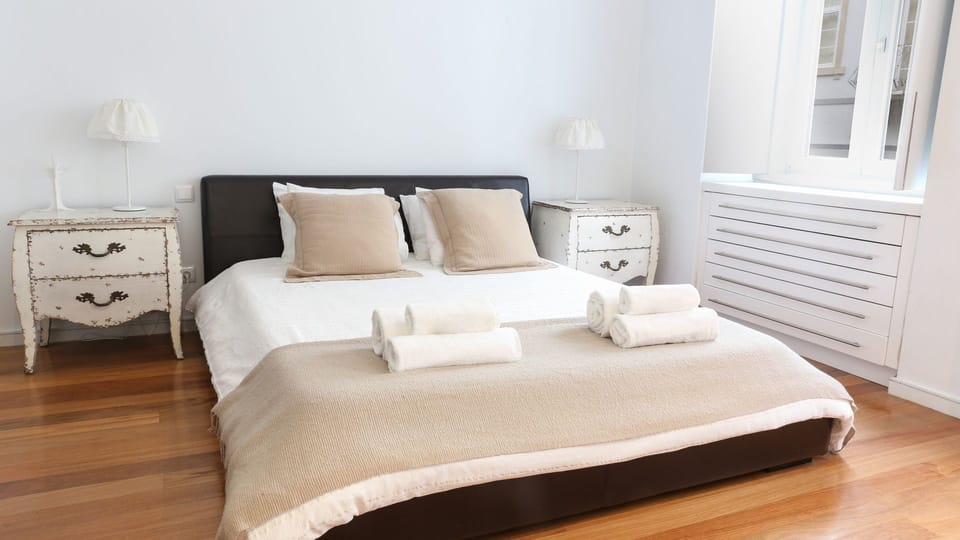 Imagine sinking into this plush queen bed after a day of exploring Lisbon, with soft lighting creating a serene atmosphere perfect for restful nights. The charming distressed nightstands add a touch of rustic elegance to your comfortable retreat. #QueenBe