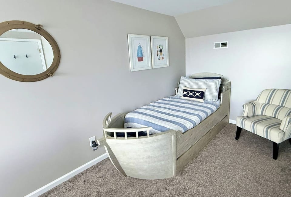 The kids will love the nautical-themed bedroom features a unique bed shaped like a boat, complemented by a round porthole-style mirror, striped accent chair, and framed coastal wall art to create a charming, sea-inspired retreat.
