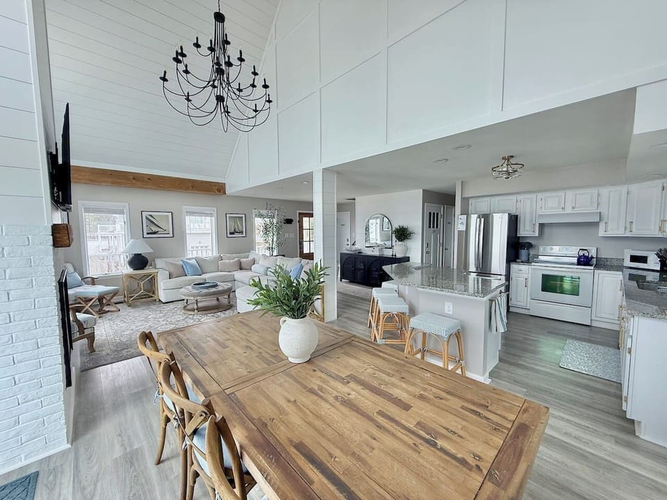 Enjoy an open-concept living space highlights a dining area with a wood table, a cozy lounge with a sofa, and a modern kitchen. A vaulted ceiling and chandelier create an airy, inviting atmosphere throughout the home.