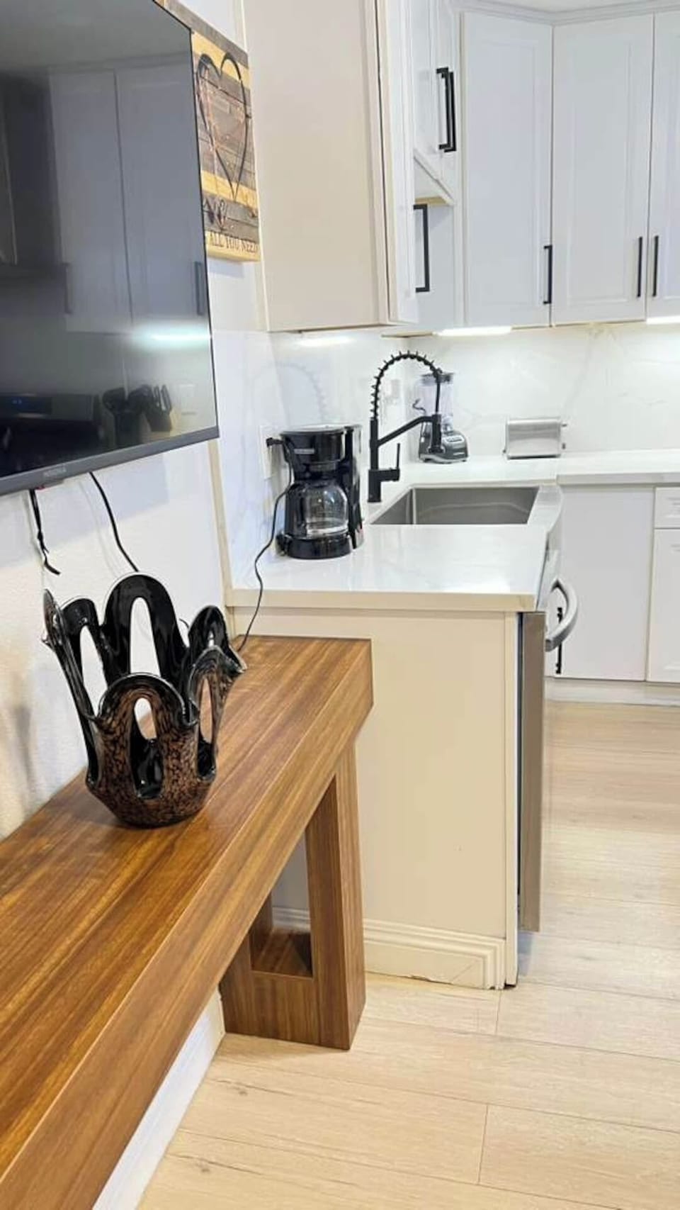 A clean, modern kitchen with sleek white cabinetry, under‑cabinet lighting, a deep stainless‑steel sink, coffee maker, toaster, and blender. Warm wood accents and organized countertop appliances create a bright, functional cooking space.
