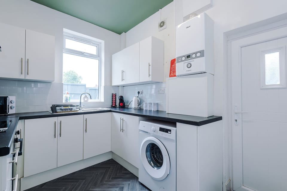 Bright, modern kitchen with white cabinets, dark countertops, and sleek appliances. Includes a washing machine, stainless steel sink, and wall-mounted boiler, a functional, well-lit space ideal for cooking and laundry convenience during your stay.