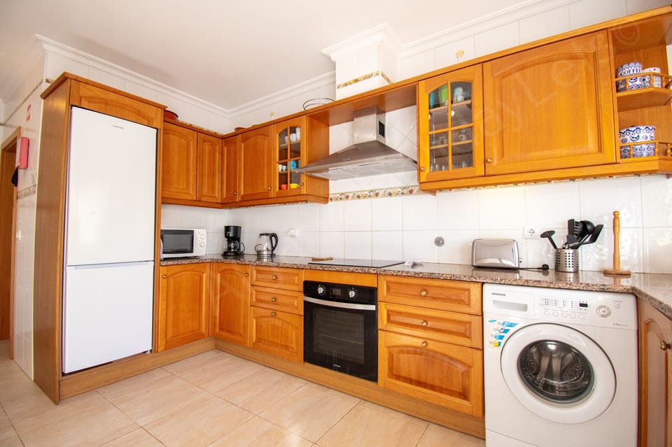Well-appointed kitchen at Casa Destino villa showing wooden cabinetry with glass-fronted cupboards, a full-size fridge-freezer, built-in oven, washing machine, microwave and kettle on granite-effect worktops, ideal for preparing meals during your self-catering holiday near Lagos, Algarve, Portugal