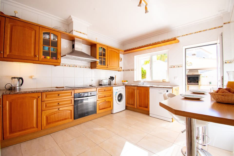 Fully equipped kitchen at Casa Destino villa with warm wooden cabinetry, glass-fronted display cupboards, built-in oven and hob, white worktops and a decorative tile border, plus a washing machine and breakfast table, providing everything you need for self-catering holidays near Lagos, Algarve, Portugal