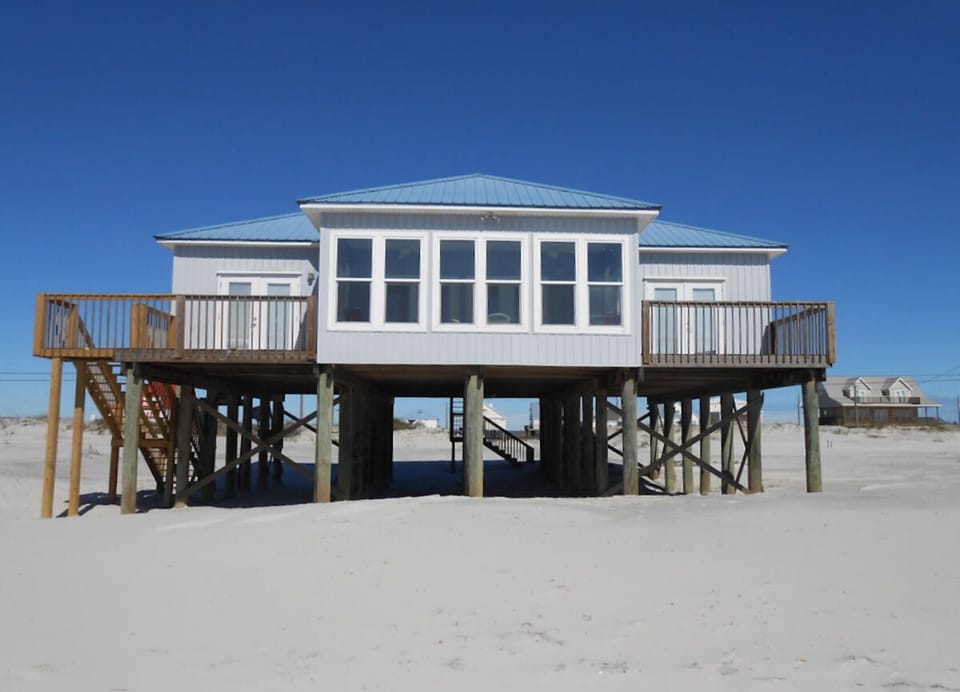 Beachfront house elevated on stilts with wraparound decks and panoramic windows facing pristine white sand and clear blue skies.