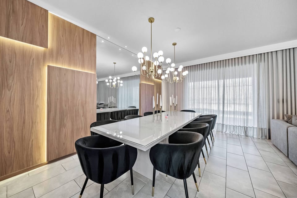 Experience dining in style at the sleek, modern table surrounded by plush black chairs, perfectly illuminated by a stunning chandelier. The warm wood accents and soft drapes create a refined atmosphere, ideal for gatherings.