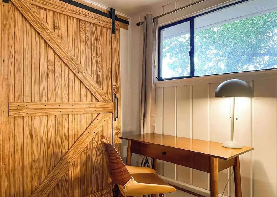 This bright bedroom features a dedicated mid-century modern desk and a stylish grey chair, ideal for remote work or creative projects. A rustic wooden barn door adds a touch of character to the space, while the large window provides plenty of natural
