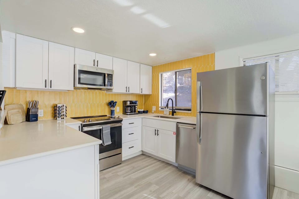 The fully stocked kitchen features stainless steel appliances, a modern gold tile backsplash, and sleek white cabinetry. It is thoughtfully equipped with everything needed to prepare home-cooked meals during your stay.