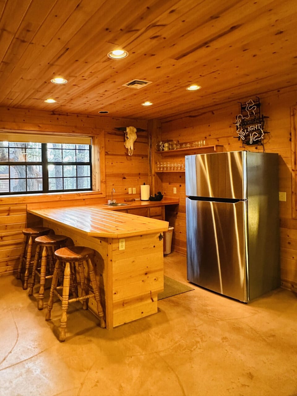 Spacious basement bar area designed for gathering and entertaining. Features generous countertop seating, plenty of cabinetry stocked with glassware for serving drinks, and a second fridge/freezer for extra storage—perfect for keeping beverages cold and e