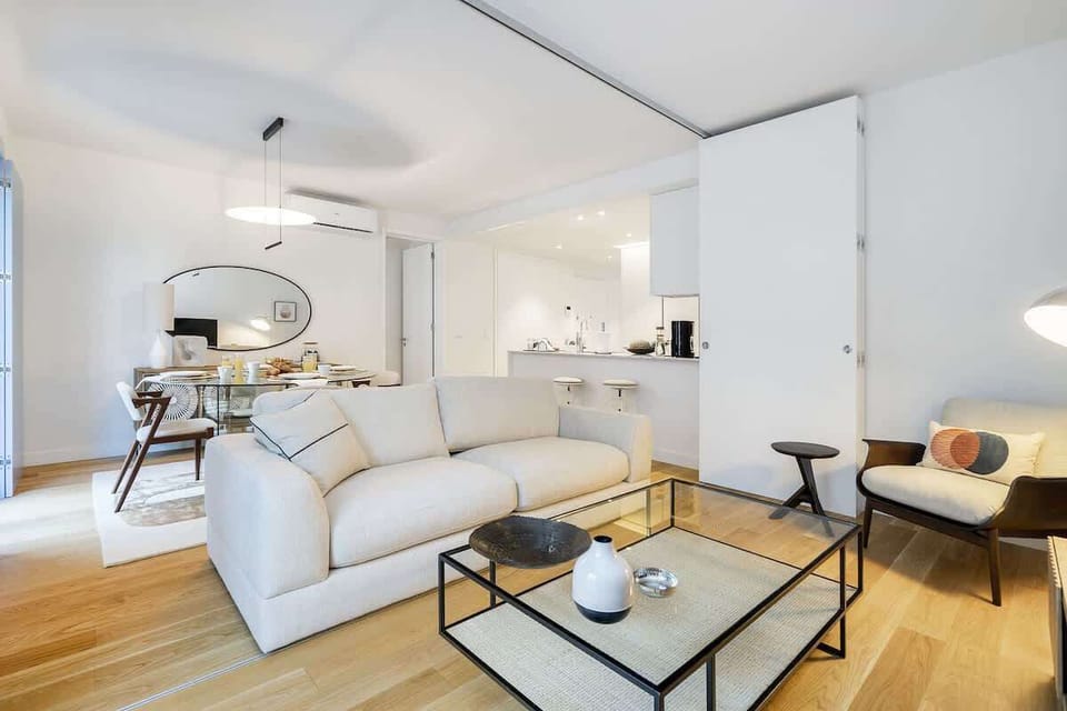 Bright and modern living room with a cozy white sofa, stylish armchairs, a sleek glass middle table. The space connects seamlessly to a dining area and kitchen.