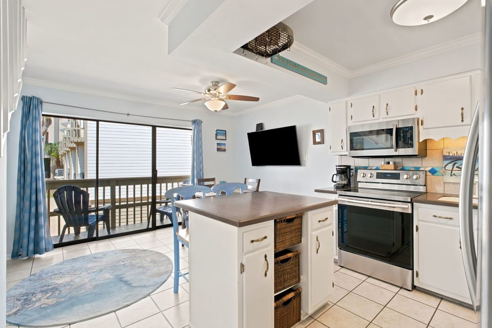 Gather around the island in this bright, coastal kitchen where cooking and conversation flow naturally. Stainless appliances, ocean-inspired accents, and a breezy balcony nearby set the stage for your beachside stay.