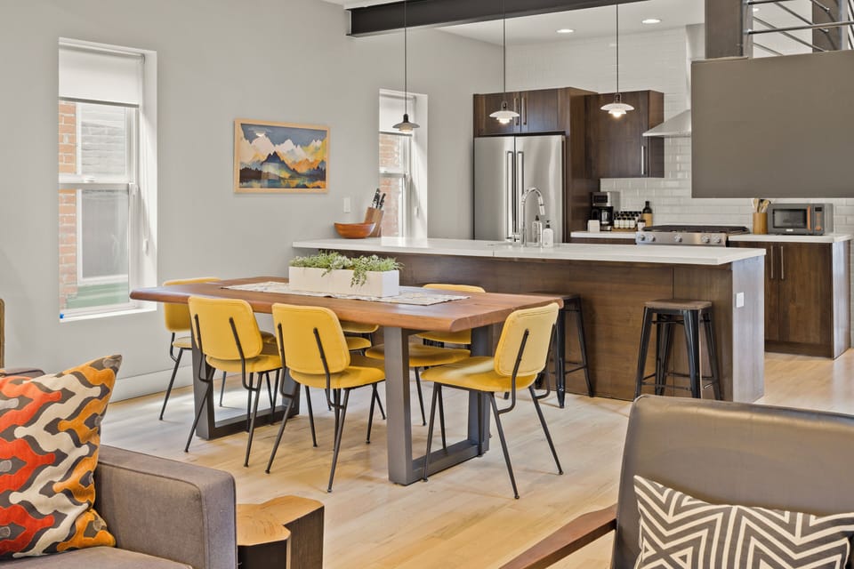 Your open-concept kitchen flows seamlessly into the dining area, where bright yellow chairs and warm wood create a cheerful space.