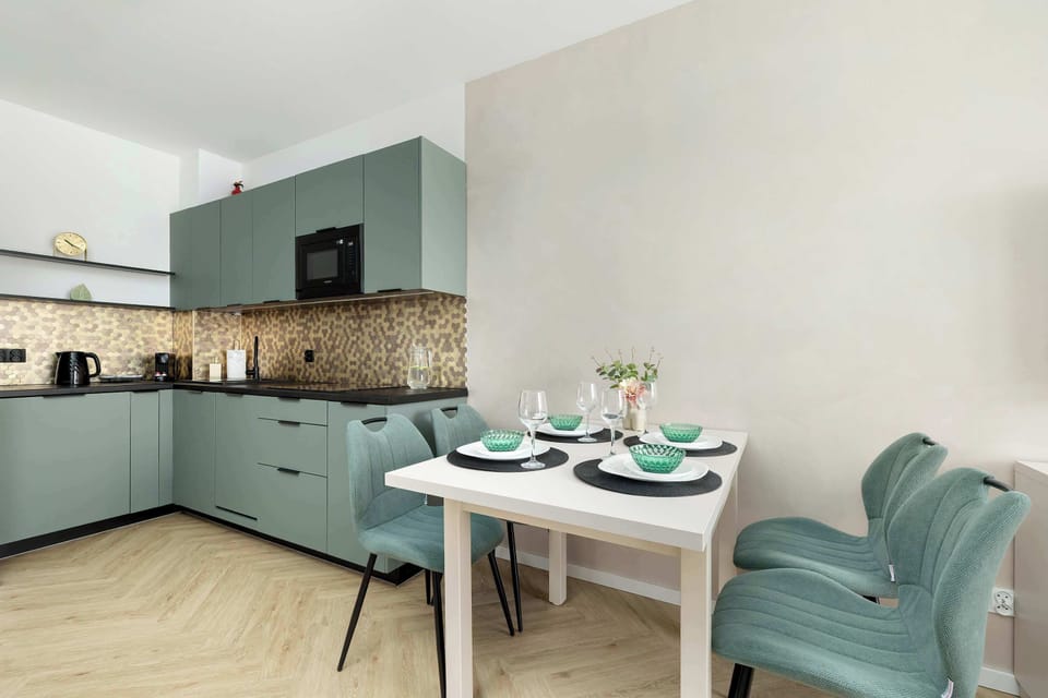A dining table set with plates and green upholstered chairs. The kitchen is visible in the background, creating an open-plan feel. The setup looks ready for everyday use.
