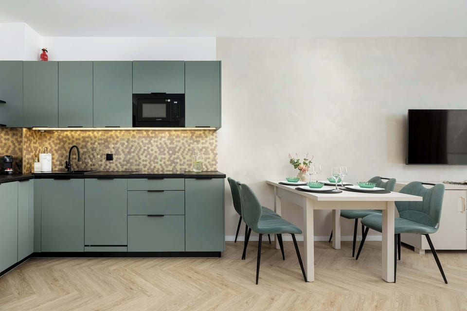 A modern kitchenette with muted green cabinets and a small dining table. Built-in appliances and patterned backsplash add character. The space feels functional and stylish.