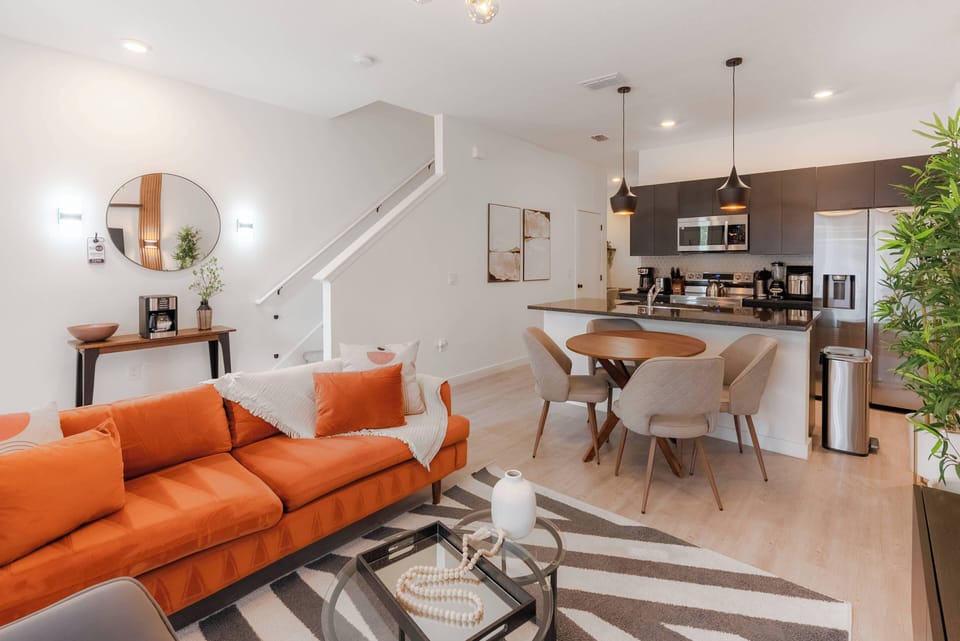 Enjoy a cozy atmosphere in this stylish living area with a vibrant orange sectional and a chic round dining table with plush chairs. The sleek kitchen complements the modern decor, enhancing your relaxation and dining experience.