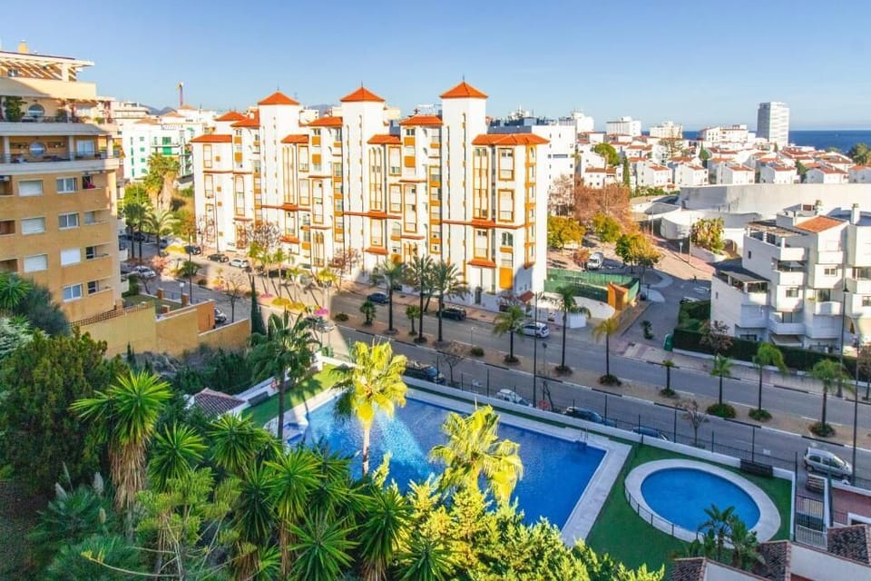 The open living area features chic decor and natural light, while the balcony offers stunning views of a sapphire pool, palm trees, and the sparkling Mediterranean sea.