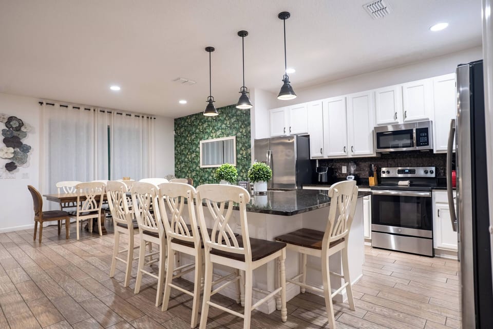 Gather in a bright open kitchen and dining area with white cabinets, stainless steel appliances, black countertops, and island seating. Pendant lights, wood-look floors, and a large dining table create a welcoming space for family meals.