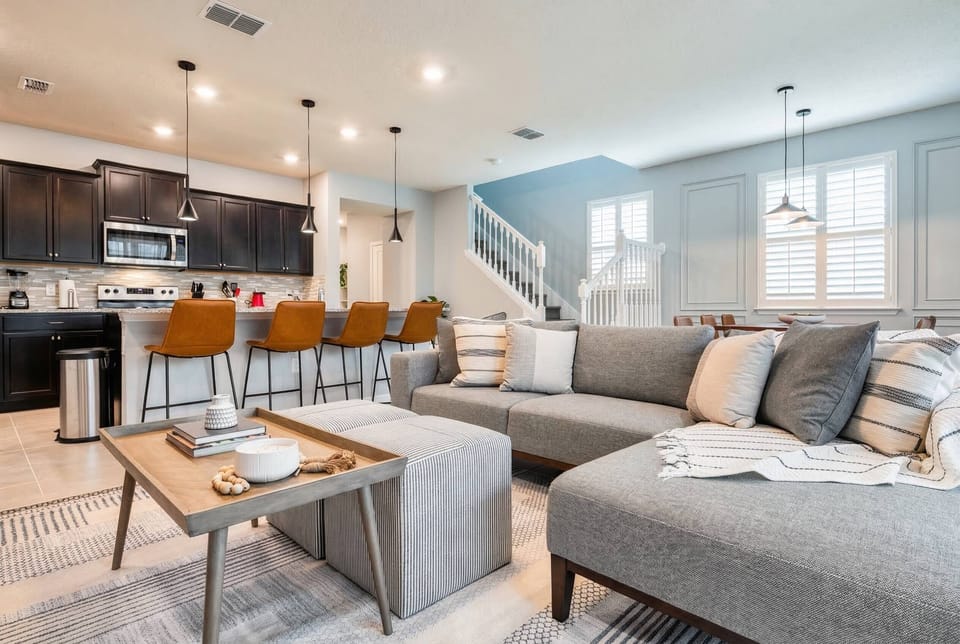 Stylish and contemporary, this living area features a cozy sectional sofa adorned with textured pillows and a chic coffee table, while the warm wooden bar stools invite you to enjoy moments in the sleek kitchen just steps away.