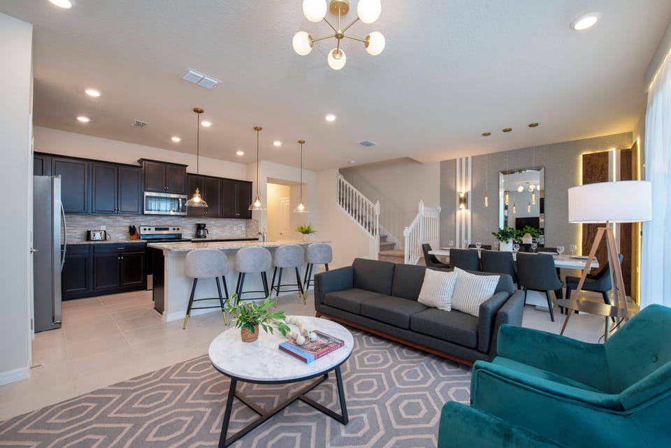 Relax in a beautifully designed living area with a cozy dark sofa and vibrant teal armchairs. Pendant lights softly illuminate the sleek kitchen, creating a warm, inviting space perfect for unwinding or socializing with friends.