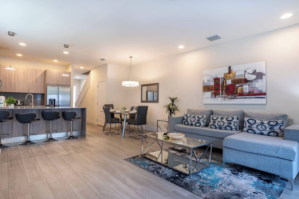 This spacious living area boasts a chic gray sofa with vibrant pillows, a warm area rug, and a sleek bar with stylish stools, creating an inviting atmosphere perfect for casual gatherings under soft, welcoming lighting.