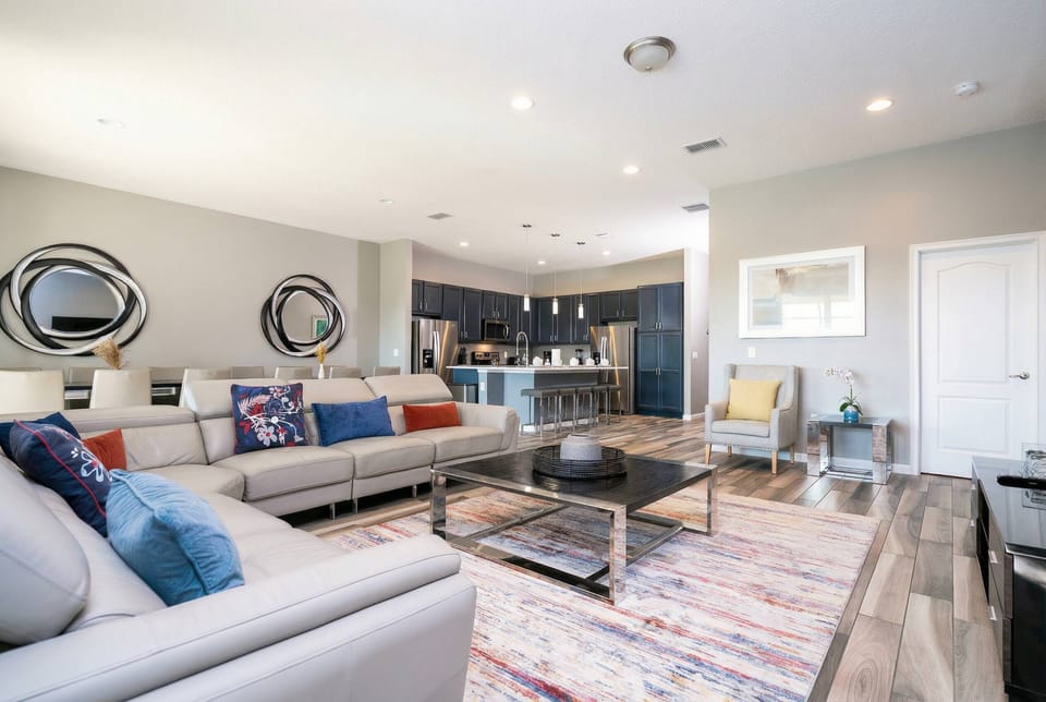 This bright and airy living area invites relaxation with its spacious sectional sofa and colorful throw pillows. Large windows flood the space with light, highlighting the rich wood flooring and open kitchen for a modern feel.