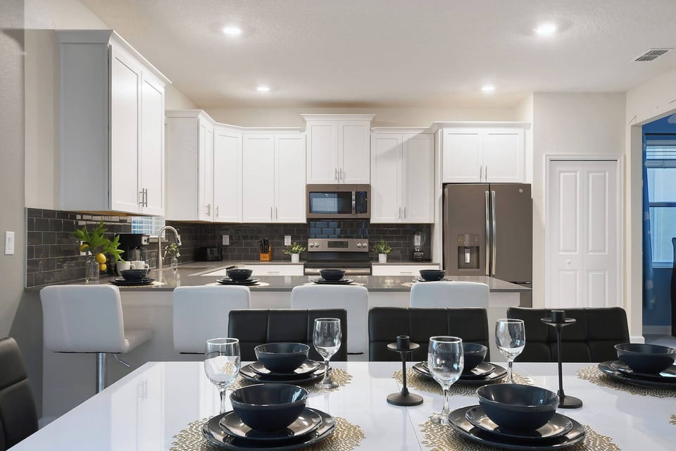 Welcome to a stylish kitchen with sleek white cabinetry and a warm gray backsplash. The modern dining table, elegantly set with black dishware, invites you to share meals and laughter in this inviting culinary space.