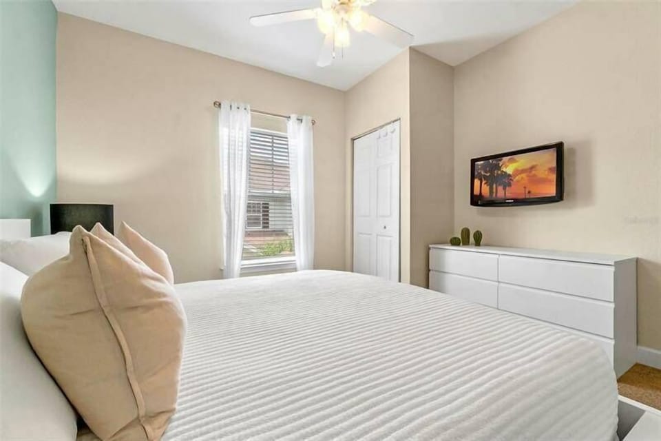 Experience restful nights in this cozy bedroom featuring soft linens on a plush bed with decorative pillows. Enjoy natural light through sheer curtains, a minimalist dresser, and a wall-mounted TV for your comfort.
