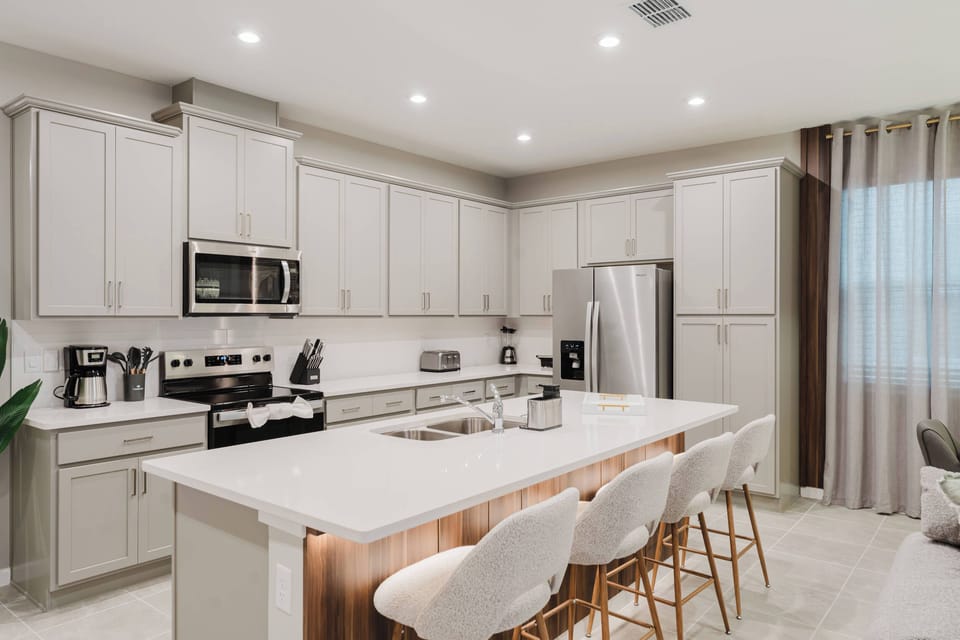 This bright kitchen boasts sleek, light-colored cabinetry and a spacious island with elegant bar stools, ideal for casual dining and socializing. Large windows fill the space with natural light, enhancing the cheerful ambiance.