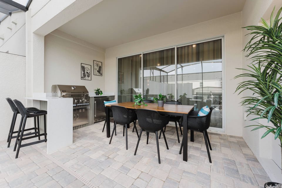 Step outside to a stylish outdoor dining area with a sleek wooden table and modern black chairs. Enjoy the built-in grill amid lush greenery and plenty of natural light from the large sliding doors.