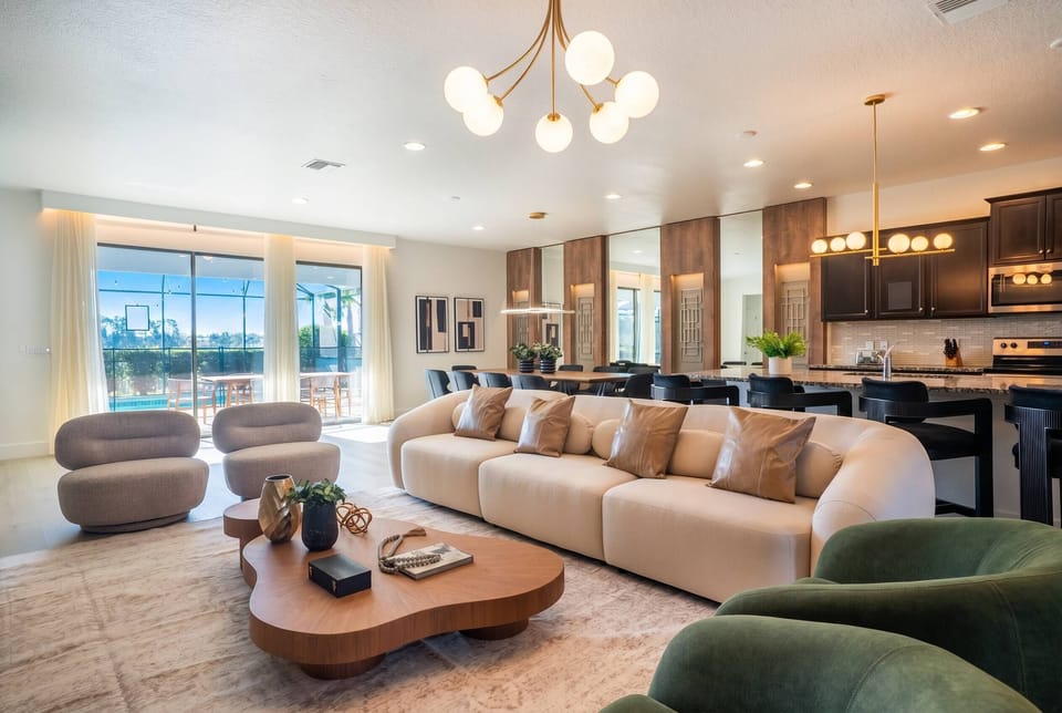 Gather in a bright open living area with a large sofa, cozy chairs, modern lighting, dining space, kitchen island, and sliding doors to the pool, creating a comfortable setting for vacation moments.