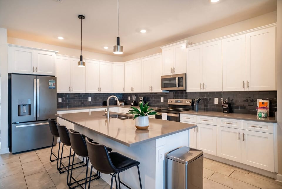 Cook and gather in a bright modern kitchen with white cabinets, stainless steel appliances, a large island, pendant lights, black bar seating, and a dark tile backsplash, perfect for family meals during vacation.