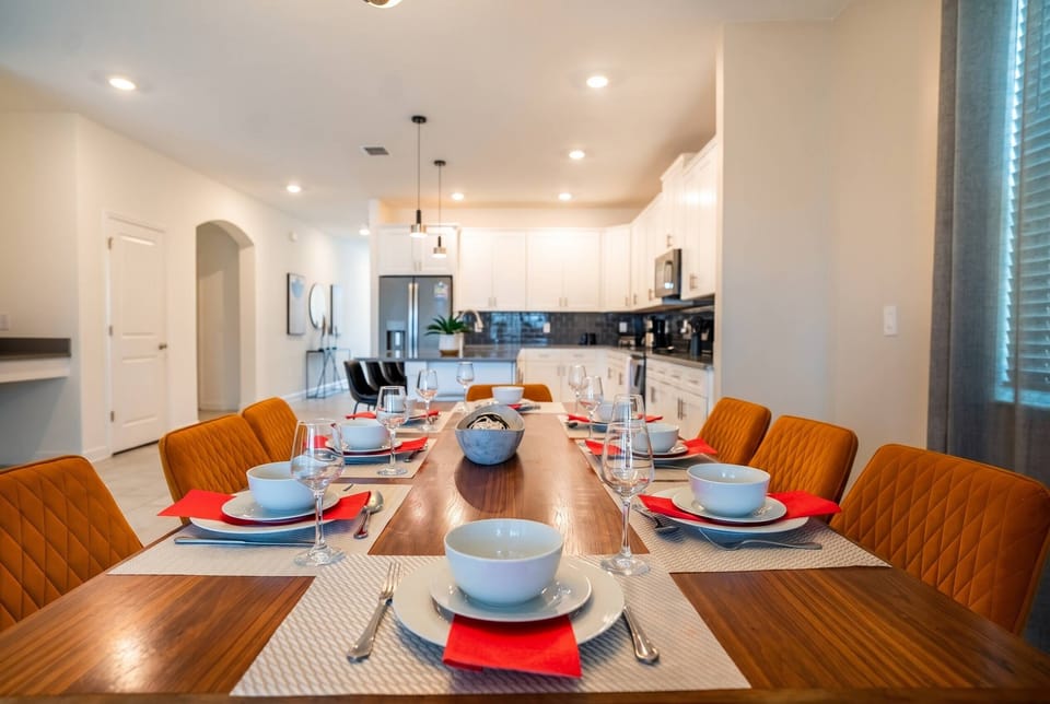 Gather in a bright dining area with a long wooden table, orange cushioned chairs, elegant place settings, warm lighting, and an open view to the kitchen, perfect for family meals during vacation.