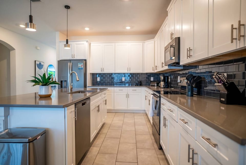Cook and gather in a bright modern kitchen with white cabinets, stainless steel appliances, a large island, pendant lights, black bar seating, and a dark tile backsplash, perfect for family meals during vacation.