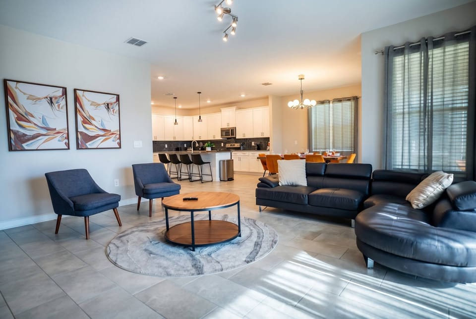 Relax in a bright living room with a black leather sectional, soft gray curtains, round coffee table, cozy accent chair, pool views, and nearby dining area, creating a comfortable space for vacation moments.