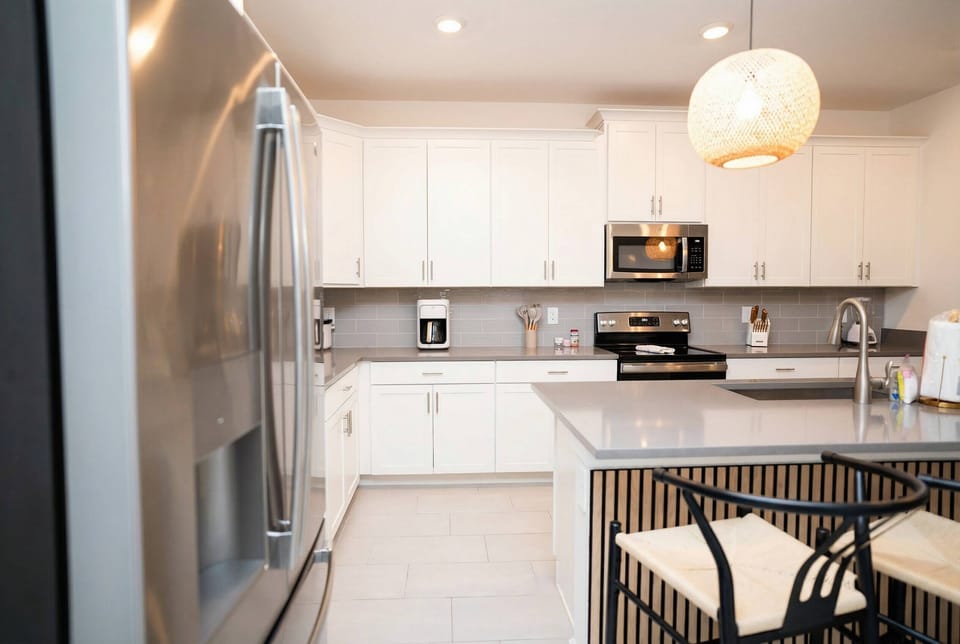 Prepare meals in a bright modern kitchen with white cabinets, stainless steel appliances, a spacious island, bar seating, soft pendant lights, and elegant gray countertops.