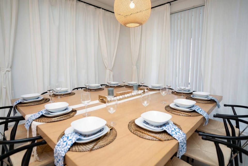 Gather around a bright dining area with a wooden table set for family meals, black chairs, soft white curtains, and a warm woven pendant light creating a cozy and elegant space.