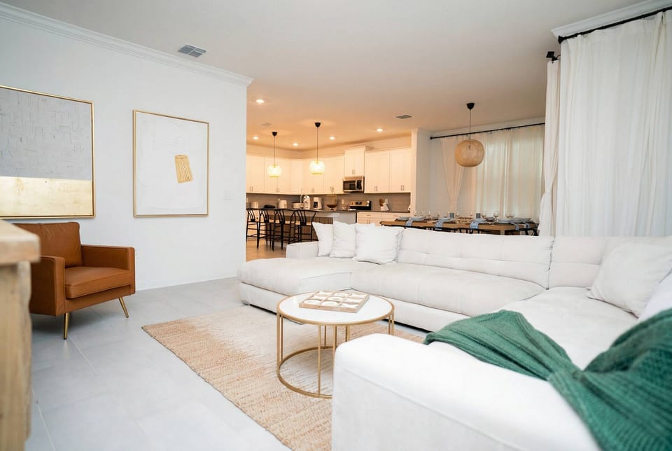 Relax in an open living area with a large white sectional, cozy accent chair, soft curtains, and warm natural decor. The bright layout flows into the dining area and kitchen, perfect for family time.