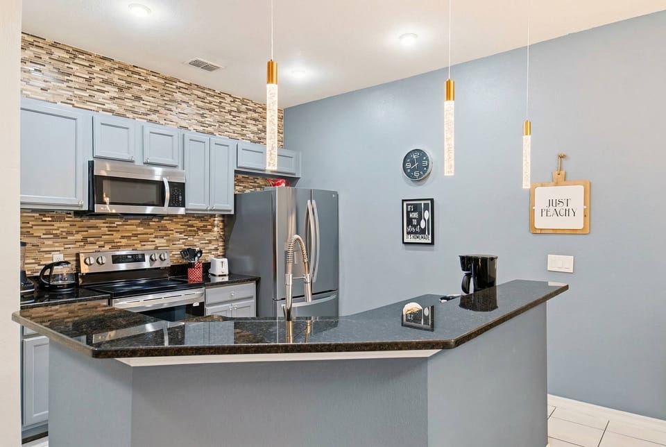 Welcome to this stylish kitchen, featuring sleek stainless steel appliances, rich stone backsplash, and glossy black countertops that inspire culinary creativity. Soft blue walls and elegant pendant lights enhance the modern vibe.