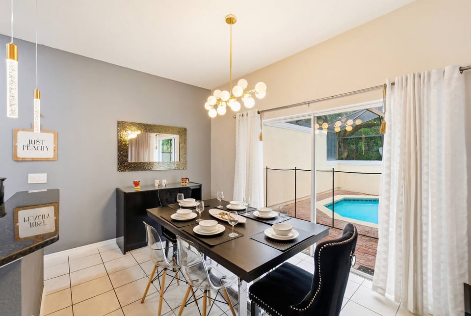 Bright and airy, this dining area features a sleek black table set for a meal, complemented by elegant acrylic chairs. Natural light floods in through the large sliding doors, revealing a glimpse of the inviting pool just outside.