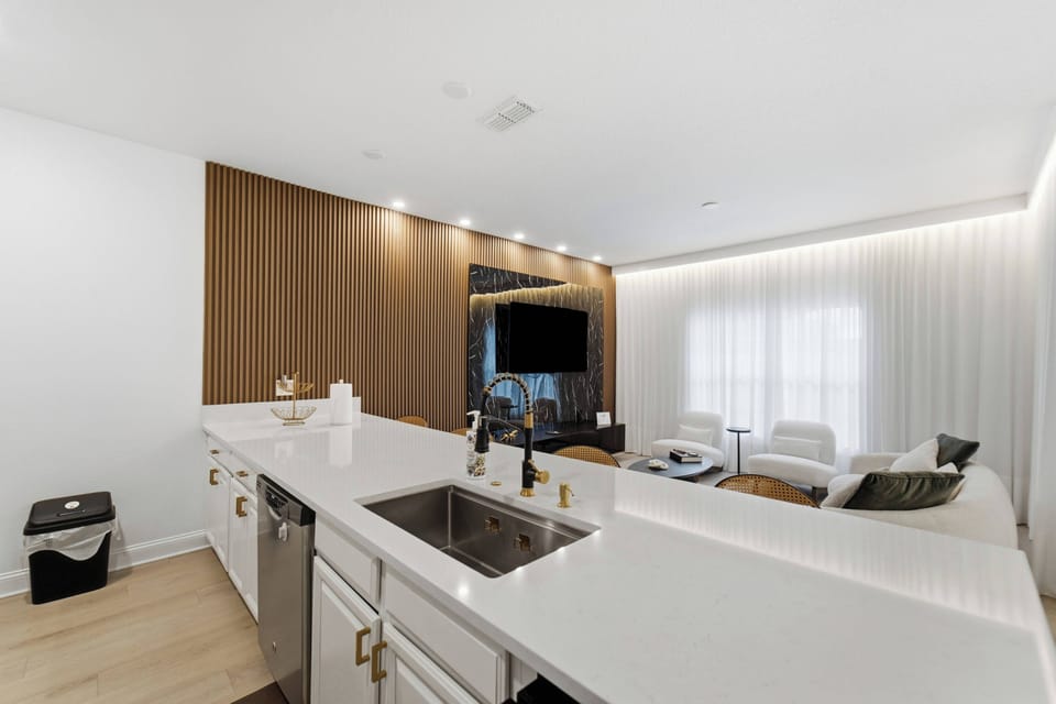 This bright, open kitchen flows into the living area, showcasing sleek white countertops and warm wooden accents. Relax on contemporary furnishings and enjoy the natural light streaming through sheer curtains.