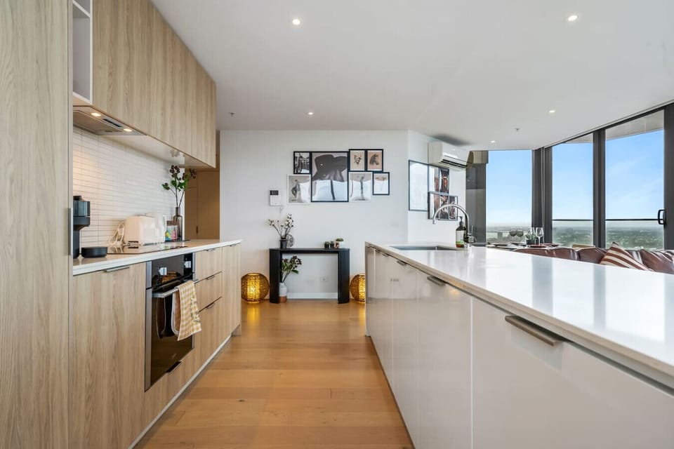 Spacious galley kitchen with warm timber cabinetry, a large island bench, oven, and Nespresso machine — flowing through to the living and dining area with floor-to-ceiling city views at the far end.