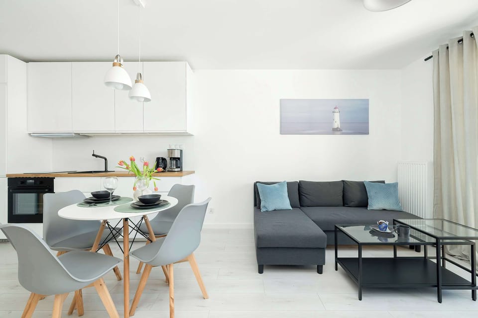 A bright living room with a sofa, coffee table, and a dining area visible in the background. The room is airy and well-lit, ideal for socializing.