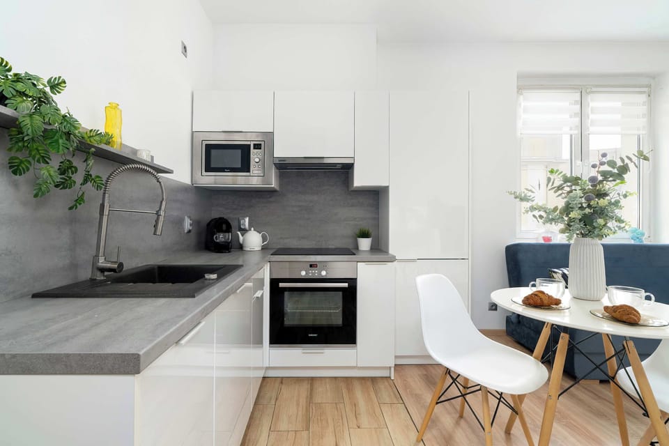 A compact kitchen with white cabinets and a gray backsplash. A small dining table sits nearby, making the space functional. Potted plants add a fresh touch.