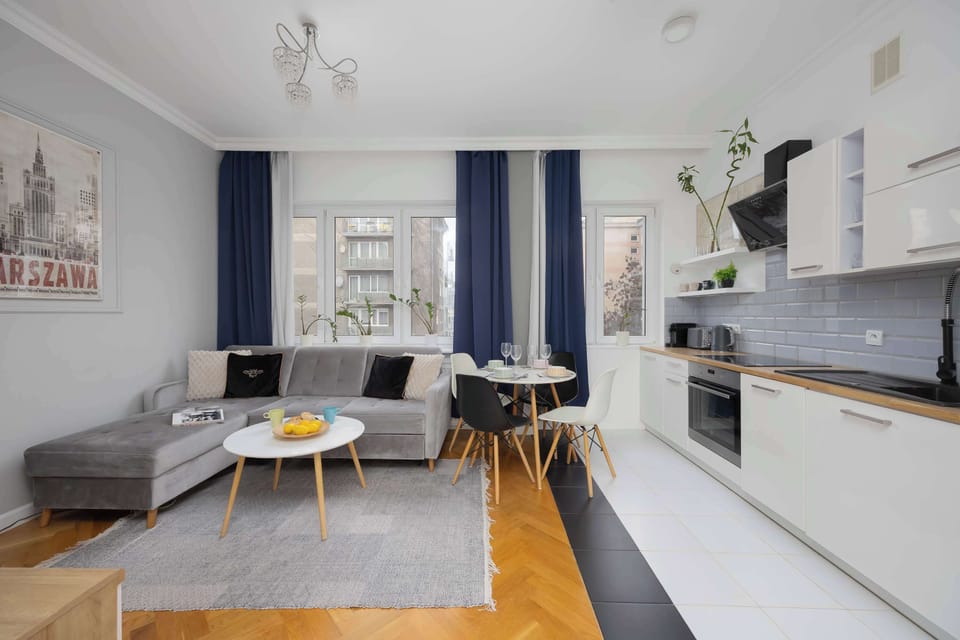 An open-plan living area combining a sofa, dining table, and kitchen. Natural light highlights the wooden flooring. The layout feels spacious and practical.