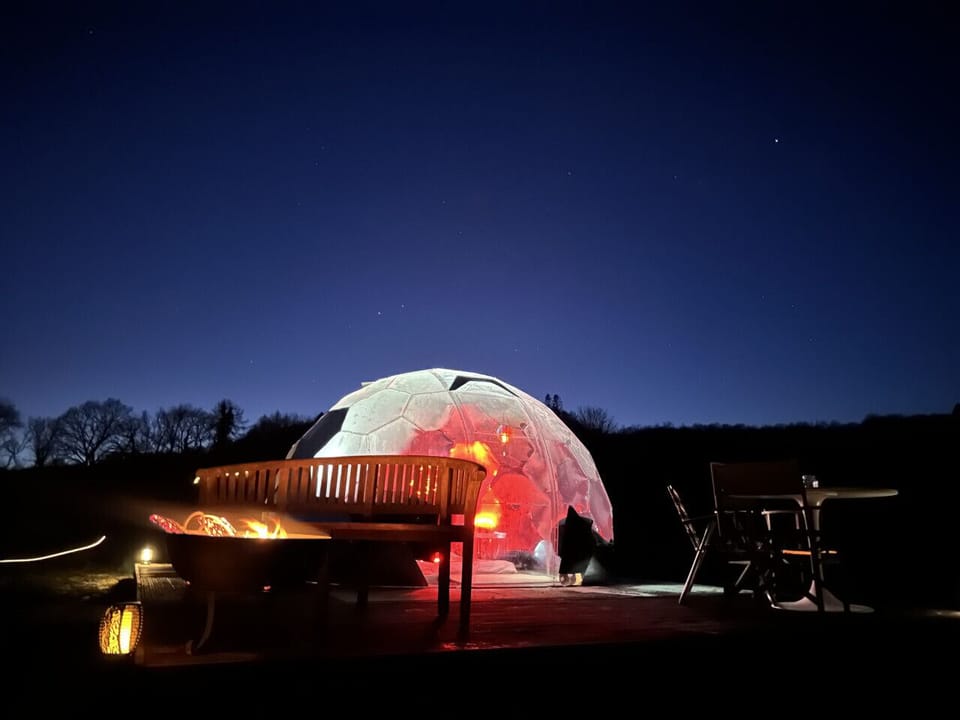 Another lit geodesic dome sits on a raised deck with outdoor seating and a fire pit under the night sky