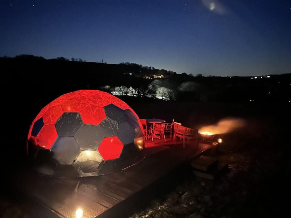 The dome glows on the deck at night with seating outside and distant lights in the background